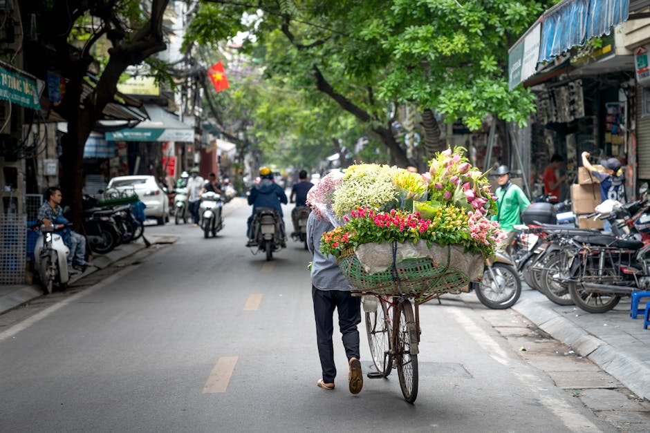 A street scene in an urban area with a flower vendor pushing a bicycle loaded with a vibrant, colorful floral arrangement, including pink, white, and yellow blooms, wrapped in light paper and secured 