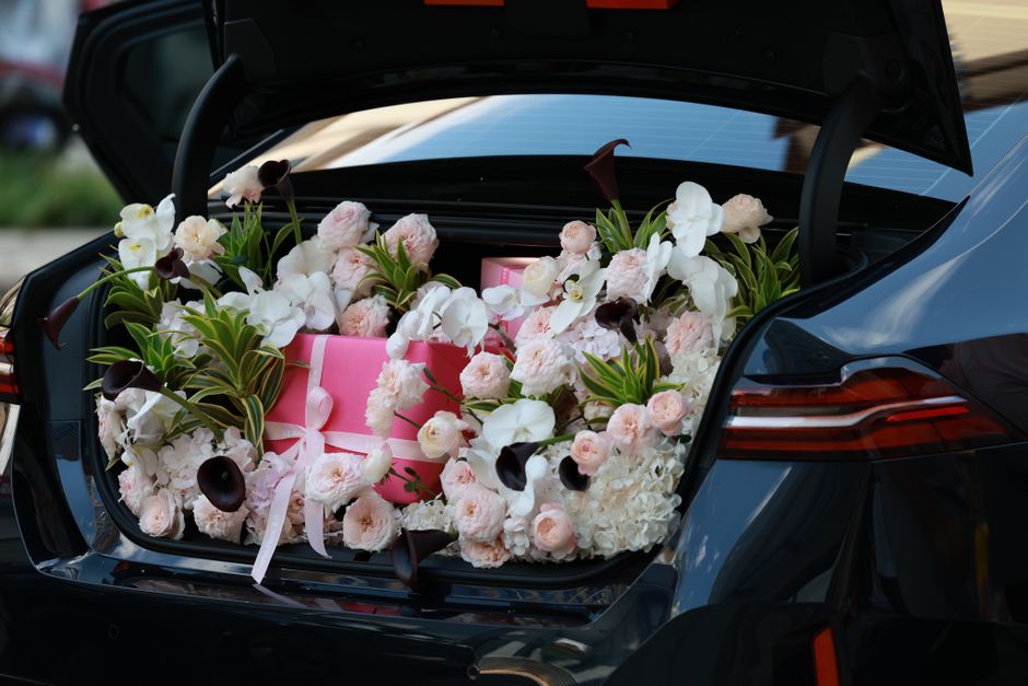 A car with its trunk open revealing a floral arrangement of fresh roses, calla lilies, and orchids in soft pink, white, and dark purple hues, surrounded by green foliage. The flowers are arranged arou
