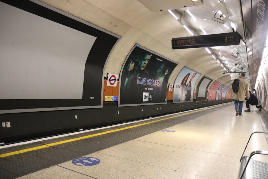 Underground station platform with curved, beige concrete walls and bright overhead lighting. Several large advertisement posters are displayed along the wall, including one promoting a TV series calle