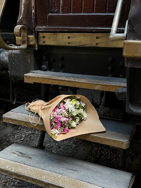 A bouquet of fresh flowers wrapped in beige paper and tied with natural twine rests on the wooden steps of a vintage railway carriage. The arrangement features white and pink blooms, with roses and sm