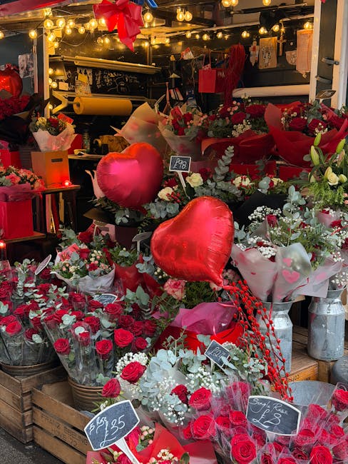 A vibrant display of floral arrangements featuring red roses, white baby's breath, and eucalyptus foliage, set in baskets and wrapped bouquets on a market stand. Bright red, heart-shaped foil balloons