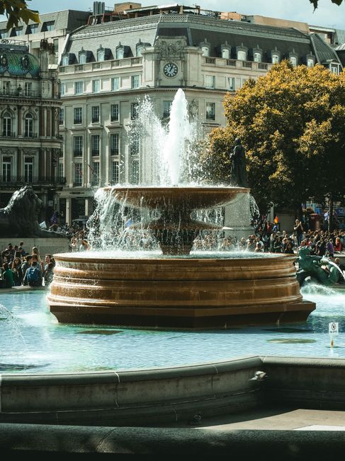 The image shows a street scene featuring Trafalgar Square in London with the famous Nelson's Column and a busy crowd around the Fountain of Nelson. In the foreground, there is a large, ornate stone fo